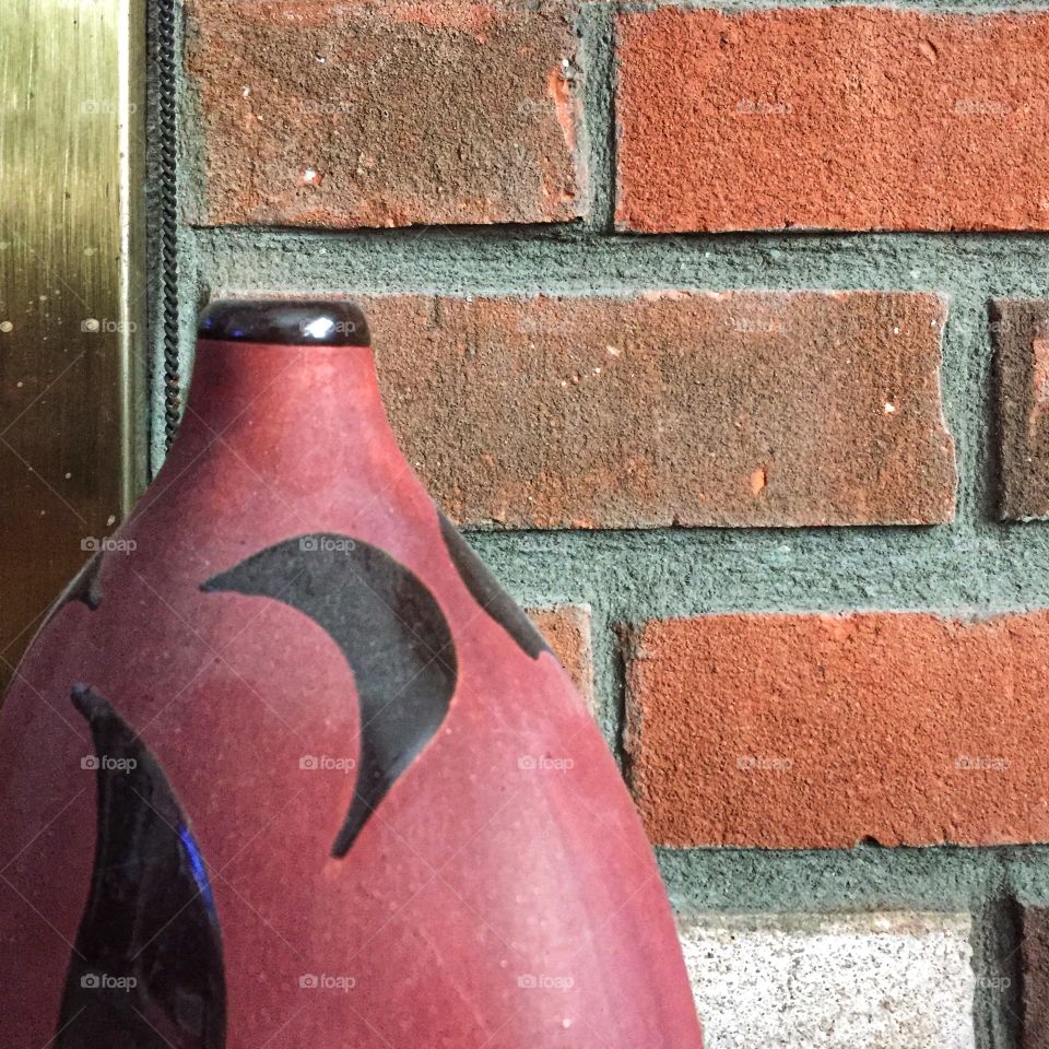 A decorative vase sits in front of a brick wall. The vase is red with black designs.