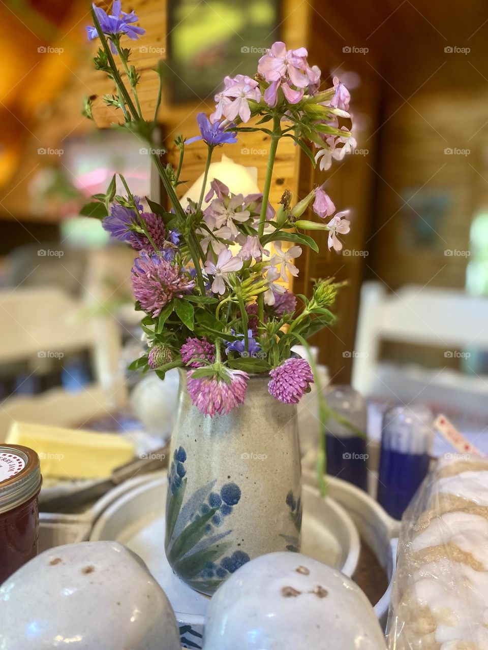 This image shows a lovely bouquet of wildflowers arranged in a ceramic vase with a blue floral design. The flowers are a mix of purple, pink, and blue hues, adding a vibrant and natural touch to the indoor cottage setting. 