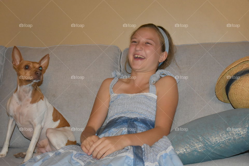 A young girl sitting on a light gray sofa in an adorable blue dress with matching head band, small dog by her side, laughs without a care. 