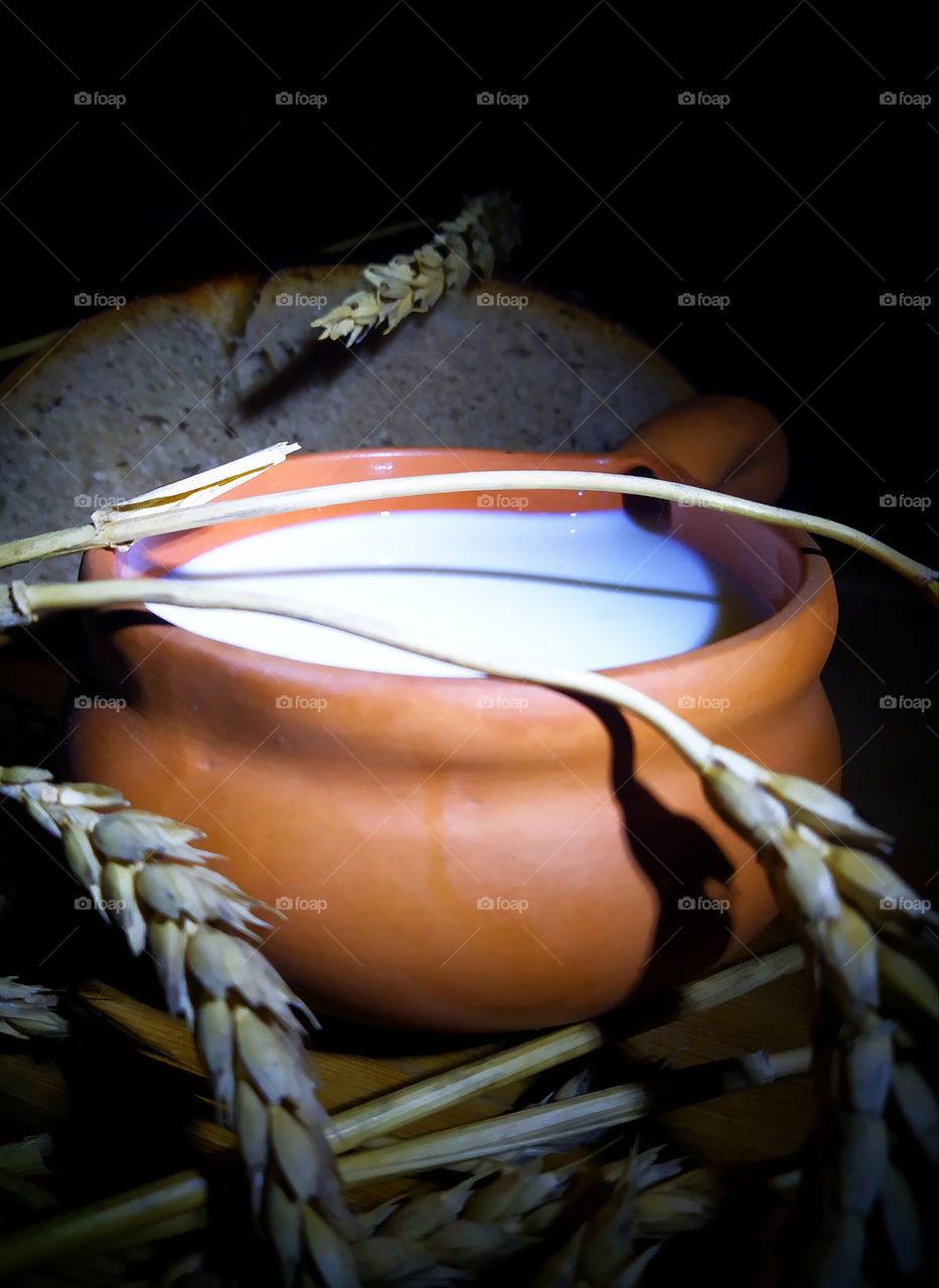 Still life: a clay mug with milk, a piece of bread and ears of wheat on a black background