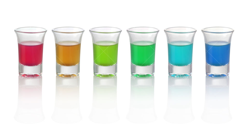 Six different colored drinks in shot glasses on white reflective background