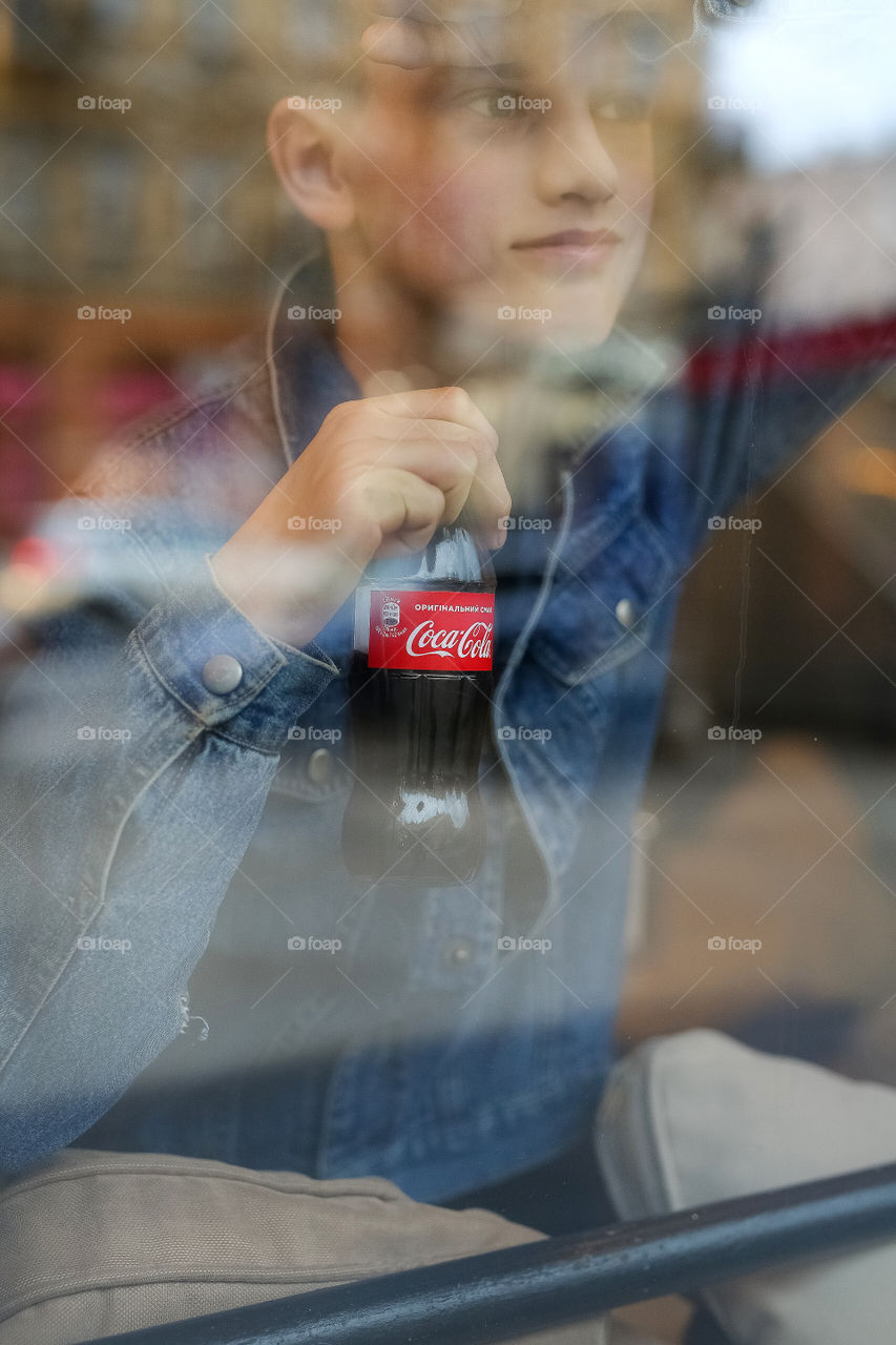 a stylish guy of twelve years old, in a denim jacket and with a bottle of Coca-Cola, stands at the window, looks, thinks, dreams.  Teenager.  generation Z