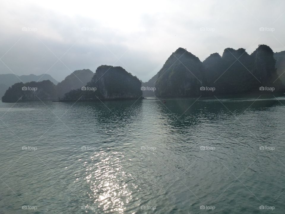 The flat blue water surface reflects the sparkling sun, many undulating mountains, cloudy sky and gray color.  the scene is magical