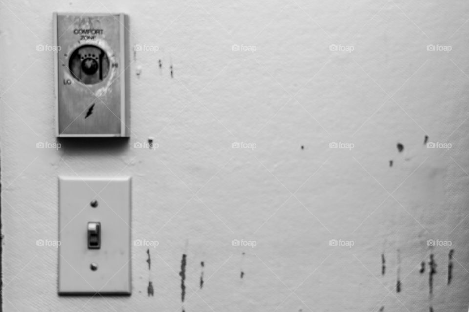 Black and white picture of neglected building wall with the ironically titled “comfort zone” thermostat placed to the side above the light switch.