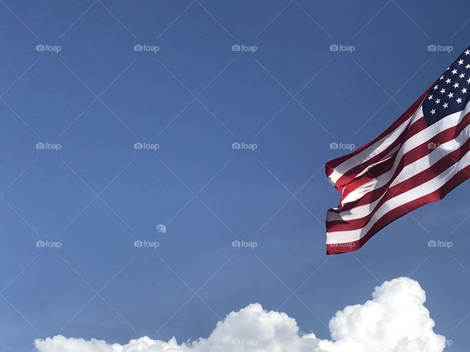 American flag waving against a blue sky and white puffy clouds. The moon is faintly in the background 
