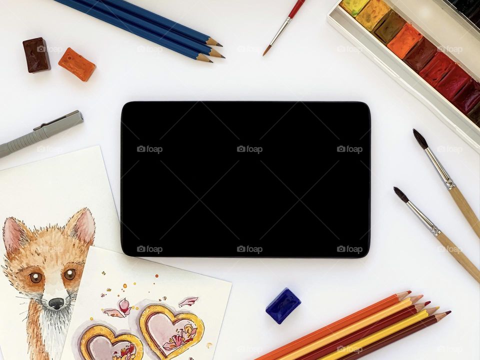Tablet with black screen, watercolor sketches, paints, brushes, colored pencils, liner on white table. Online education.