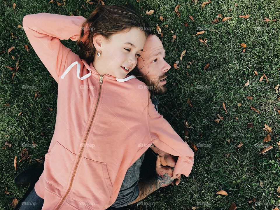 Cute little girl laying on top of her dad in the grass at the park with a sweatshirt on and looking so happy. 