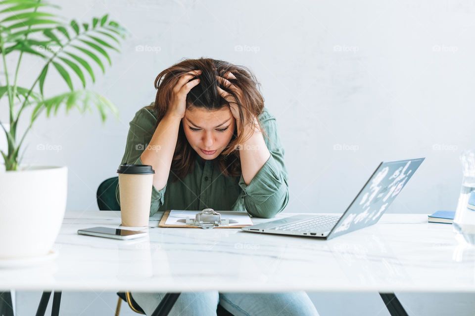 Young thinking unhappy brunette woman plus size working at laptop on table with house plant in bright modern office