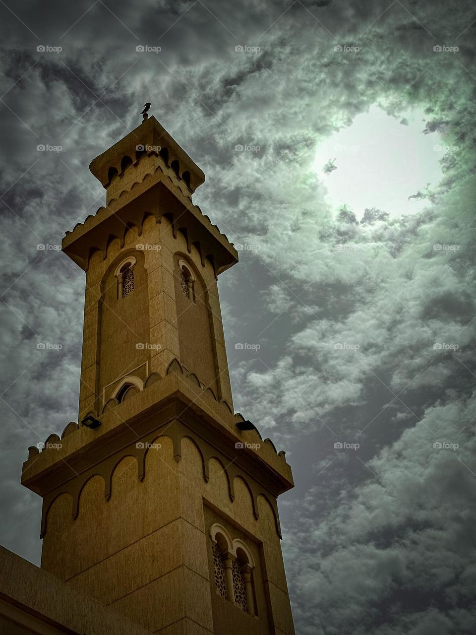 A low angle shot of a mosque minaret with cloudy skies concealing the sun 