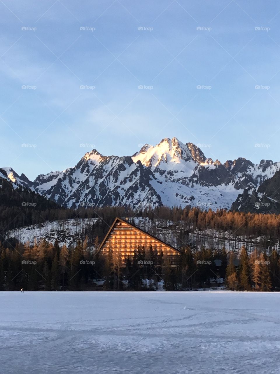 Snow capped mountain top has a sun reflection on the cabin and the trees. A blue sky is the background