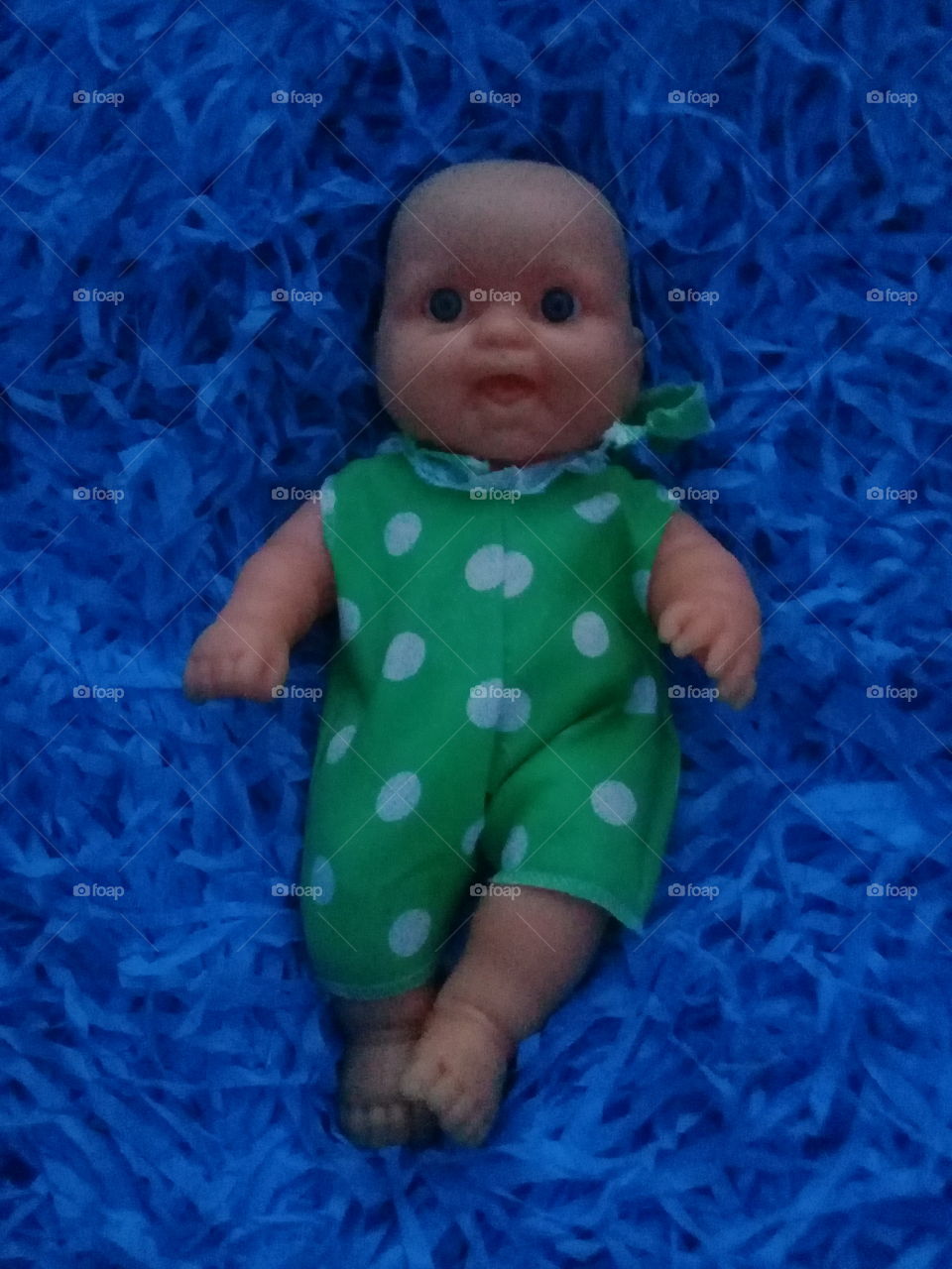 muñeca de goma acostada sobre una cama de paja azul y vestida con un enterito de tela verde.