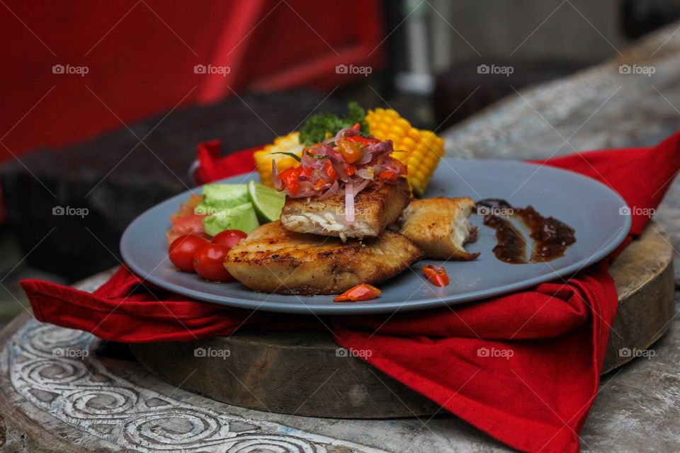 Tuna steak cooked with corn garnished with cherries, sliced tomatoes and lemon topped with sliced hot chili peppers on a baby blue melamine plate lined with red cloth and thick wood. Restaurant food.