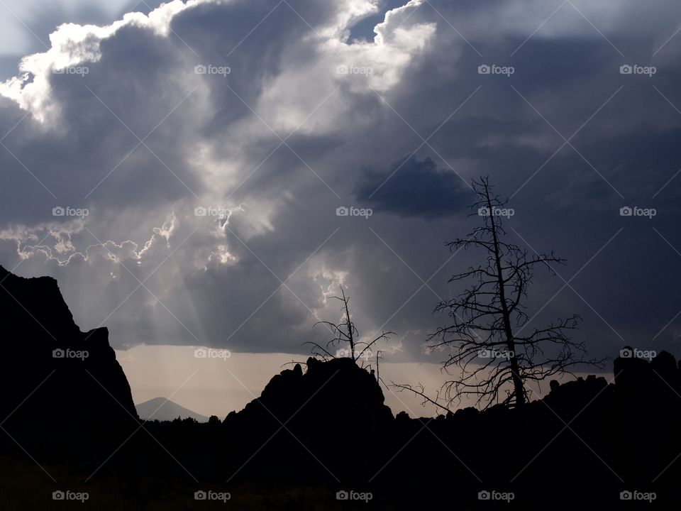 Beautiful sun rays break through the clouds after an evening summer rain storm at Smith Rocks in Central Oregon with Black Butte in the background and silhouetted trees in the foreground.
