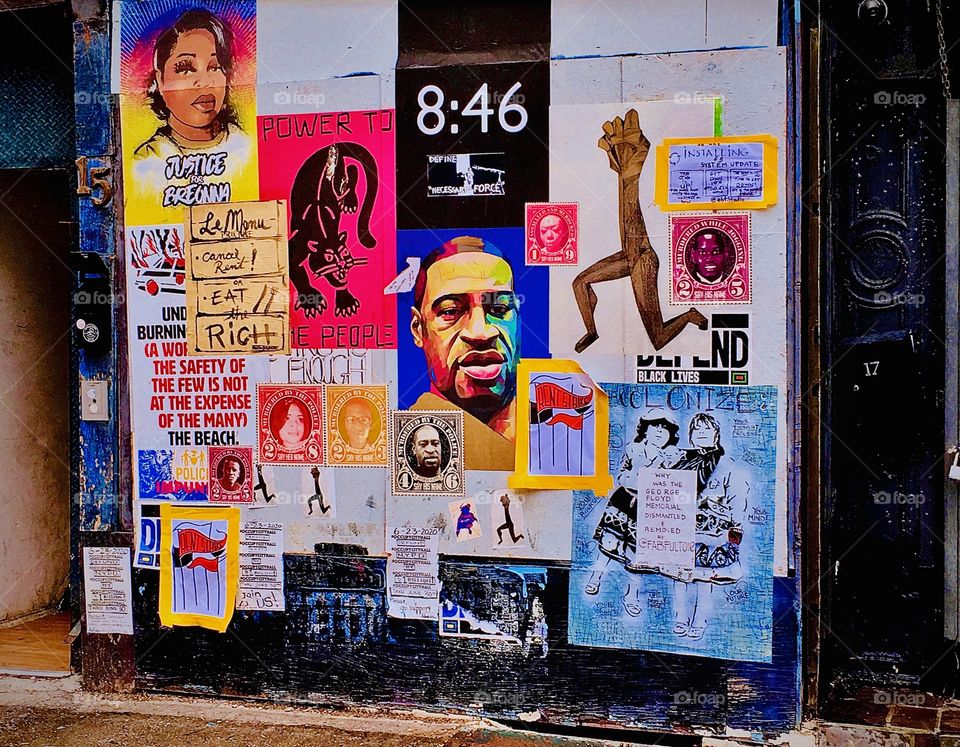 A mural collage put together from multiple flyers as well as works of art and graphic design in print is located next to a plaza with benches, umbrellas, outdoor tables and chairs on Fulton Street in Forest Green of Brooklyn, New York. Photo 2020