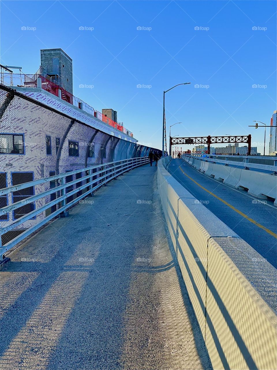This is the pedestrian lane of the „Pulaski Bridge“ at „Newtown Creek“ that connects „Greenpoint“, Bklyn to LIC, Queens in the direction we are currently heading. The evening sun illuminates the walkway and casts shadows. 2024. Hypnotic Productions