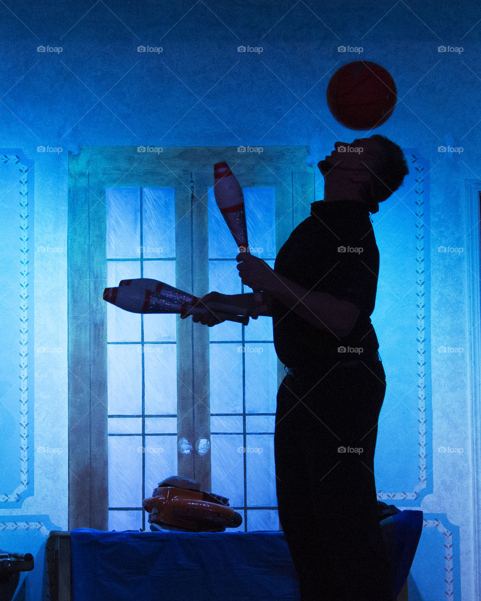 Man juggling bowling pins and also a basketball on his head, silhouette with blue background 