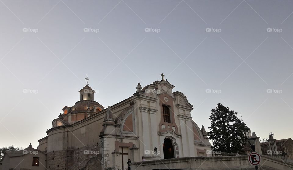 iglesia deAlta Gracia con  cruces de día y cielo celeste con árboles y parde d piedra