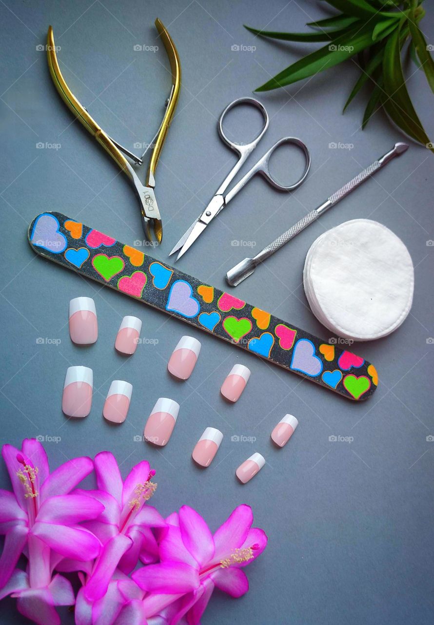 Manicure. The gray background is divided by a multicolored nail file. On one side are false nails and three pink cactus flowers, on the other side are: metal nail scissors, cuticle nippers, spatula, cotton pads and green leaves
