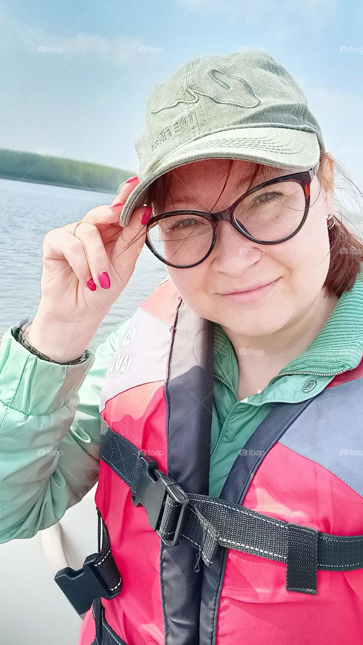 a woman in a baseball cap, a boat trip, a woman in a life jacket, a sunny day, a pond, water, a blue sky, a boat, a portrait, a girl with glasses