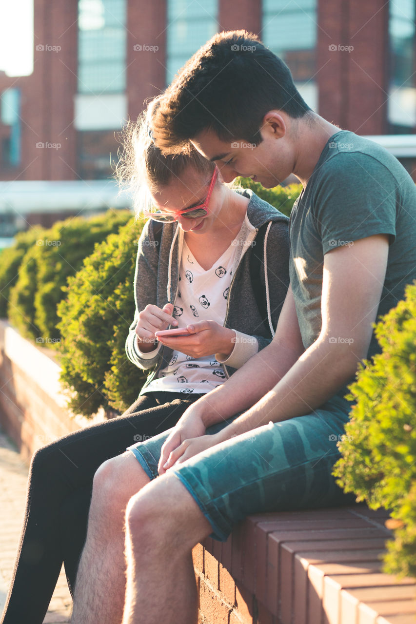 Couple of friends, teenage girl and boy, having fun together, using smartphones, sitting in center of town, spending time together. Real people, authentic situations