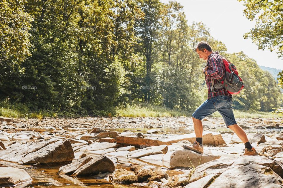 Trekking with backpack concept image. Backpacker wearing trekking boots crossing mountain river. Man hiking in mountains during summer trip. Vacation trip close to nature. Natural scenery