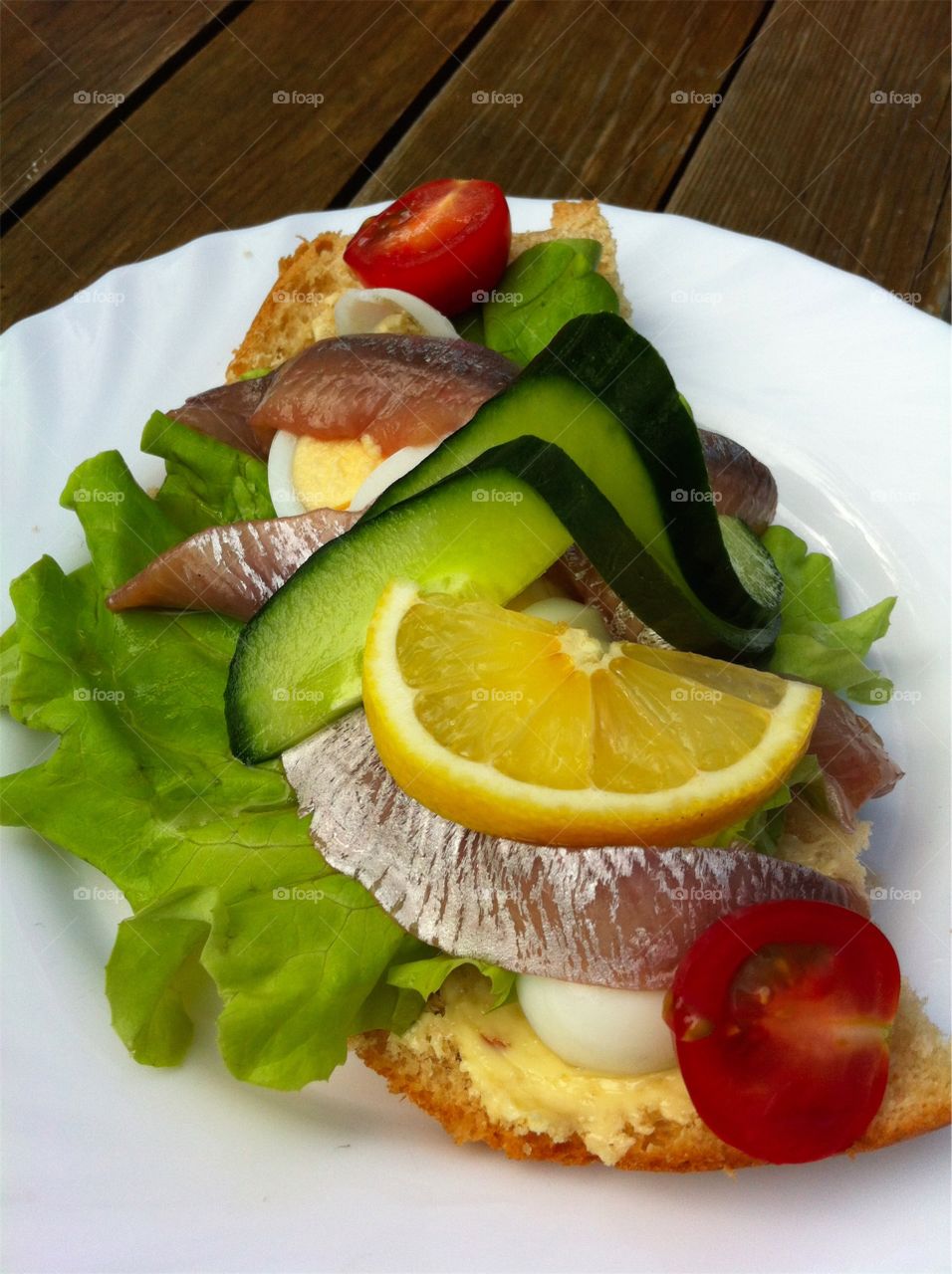 Sandwich with egg and anchovies and vegetables on white plate on wooden table outdoors.