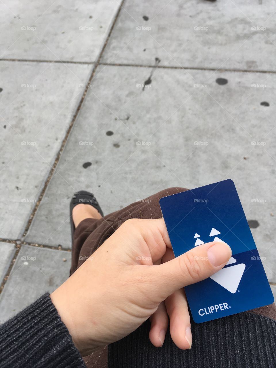 Waiting for a bus holding a blue clipper bus card. Downwards angle with knee, leg, and sidewalk
