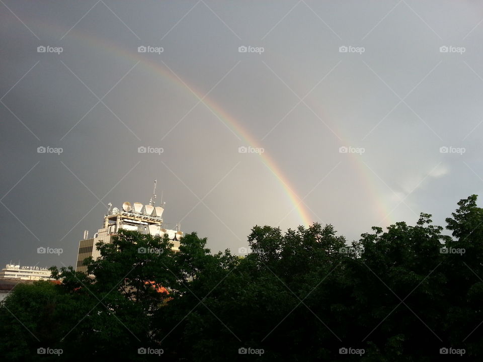 Two rainbows shine through a dark grey sky behind dark trees. In the background are two city-esque builings that seem to be leaning towards the rainbows.