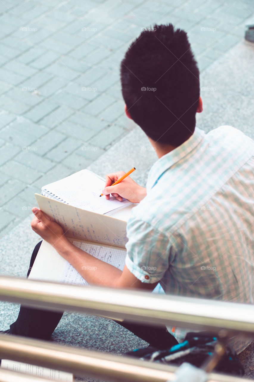 Student making a notes sitting on stairs outside of university. Young boy wearing blue shirt and dark jeans