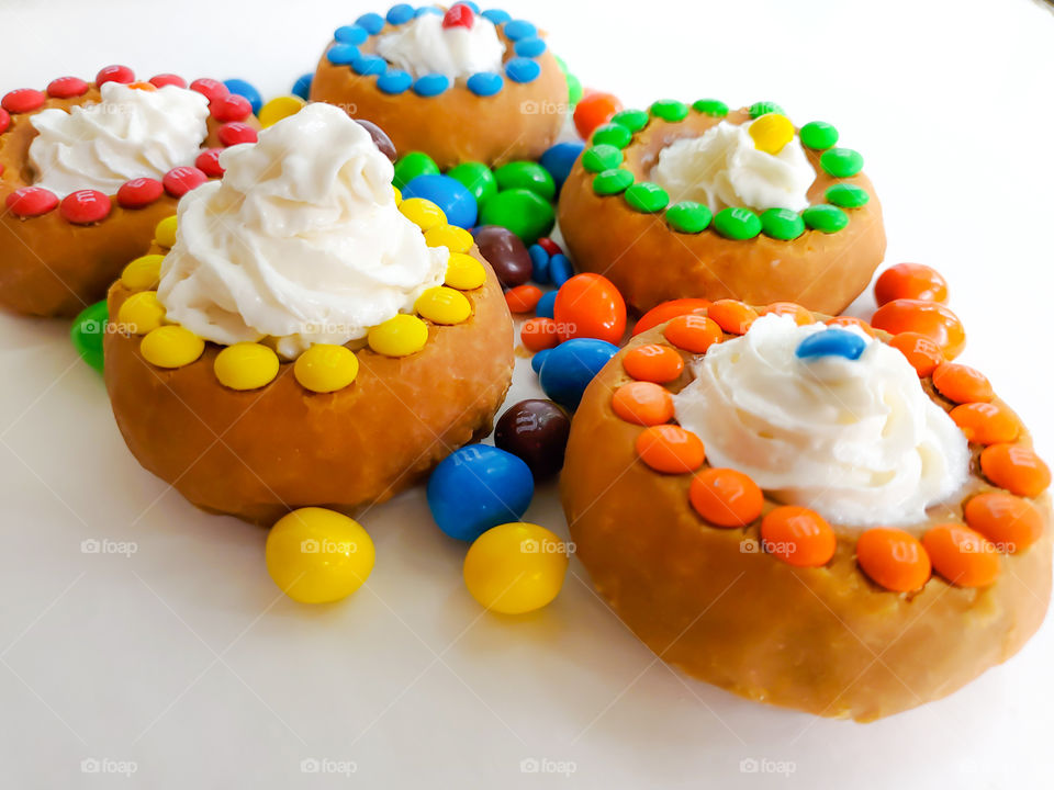 Donuts decorated with M&M minis, topped off with whip cream and surrounded by peanut M&Ms on a white surface.