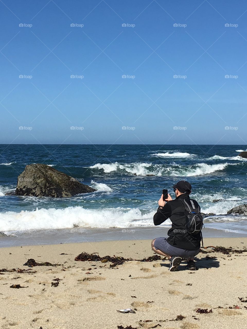 Male hiker kneeling down on a beach taking a photo of the ocean waves.