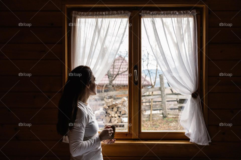 Woman drinking a hot cup of tea while looking out the window of a mountain cabin, with a view towards the mountains.
