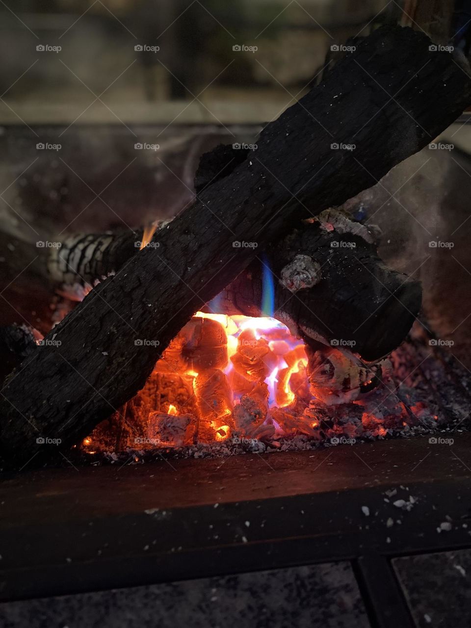 This is a photo of a campfire with burning logs. The embers and flames show a mix of red, orange, and blue colors, indicating the high temperature and presence of different chemicals.