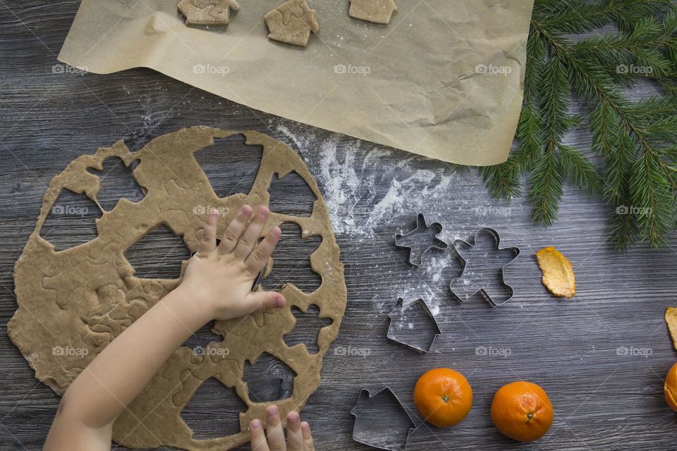 Little child helps cooking christmas ginger cookies on a wooden table with tangerines and green Christmas trees.