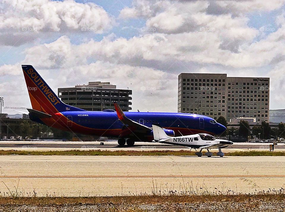 A small airplane is seen taxiing next to a big commercial airliner on the ground at an airport, with buildings in the background. The airplane is a Boeing 737 and the small plane is a Sling 2 light sport aircraft. 