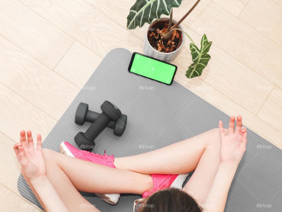 One caucasian teenage girl sits on a gray mat with a flower, two black dumbbells, a smartphone with a green screen and meditates with her legs and fingers crossed, flat lay close-up.