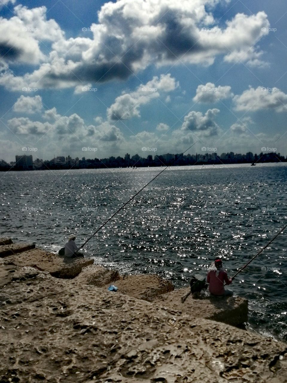 Some men practice a hobby fishing on the scenic Sea of Alexandria with apartment buildings, clear blue skies and beautiful clouds