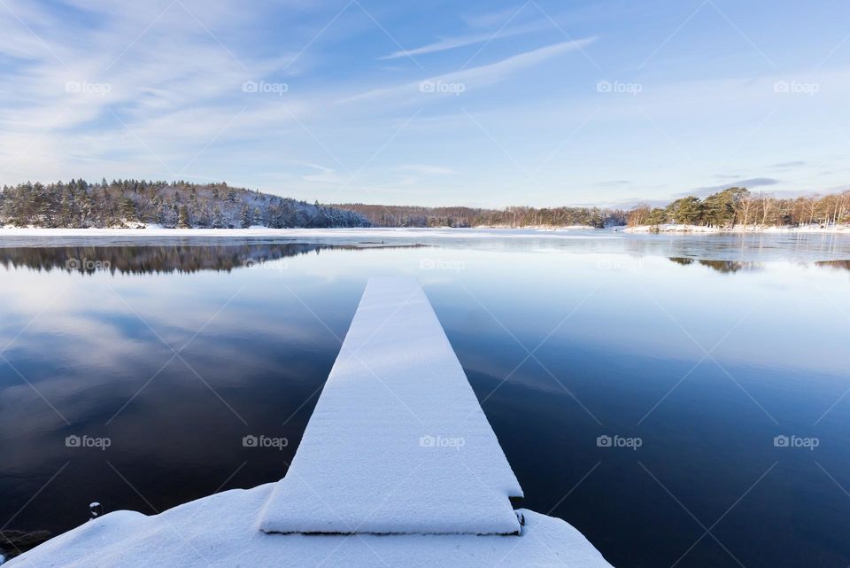 Peaceful winter landscape, snow covered jetty in partly frozen lake with sky reflection 