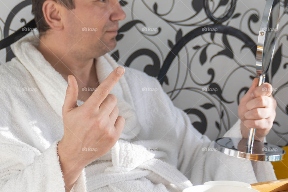 A man and home spa care at home takes care of himself and smears his face with cream, does a manicure at home on a blue background and with oranges and rests.