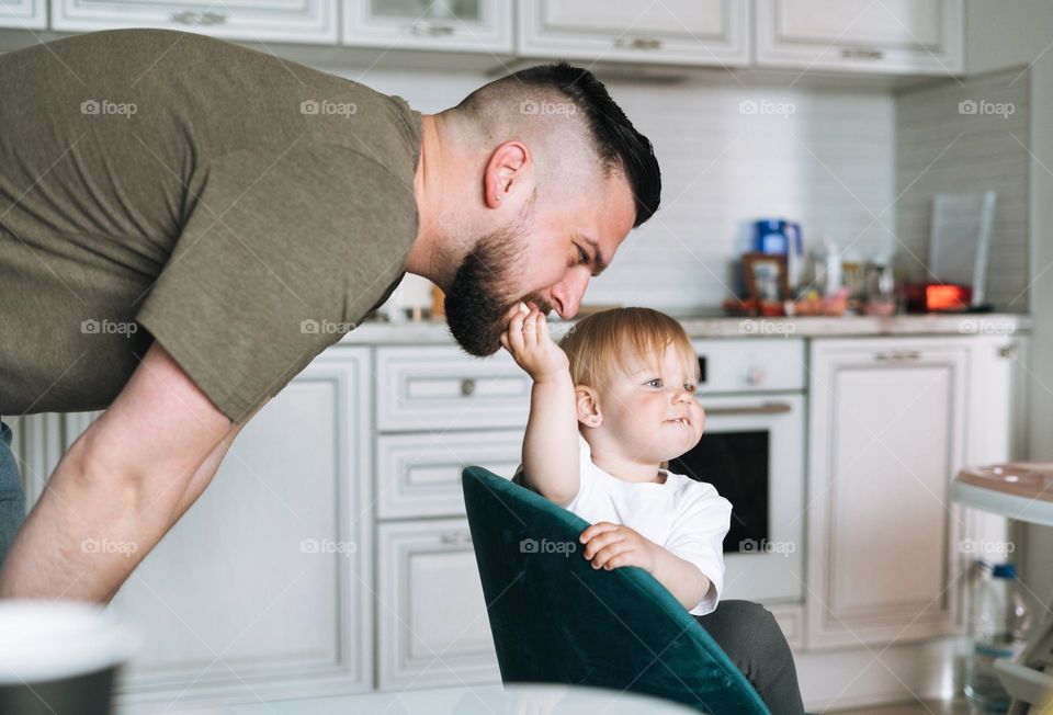 Happy father young man feeding and having fun with baby girl little daughter in kitchen at home