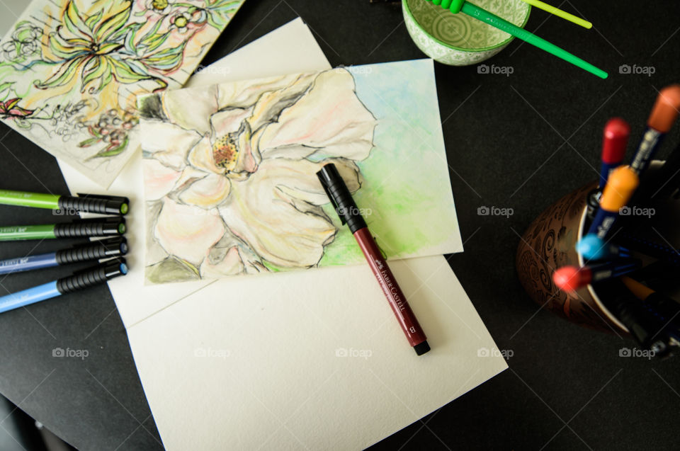 Paintbrush, water and Faber-Castell PITT artist pens on desk with watercolor style painting sketch using Faber-Castell pens high angle view of spring flowers on sketch books