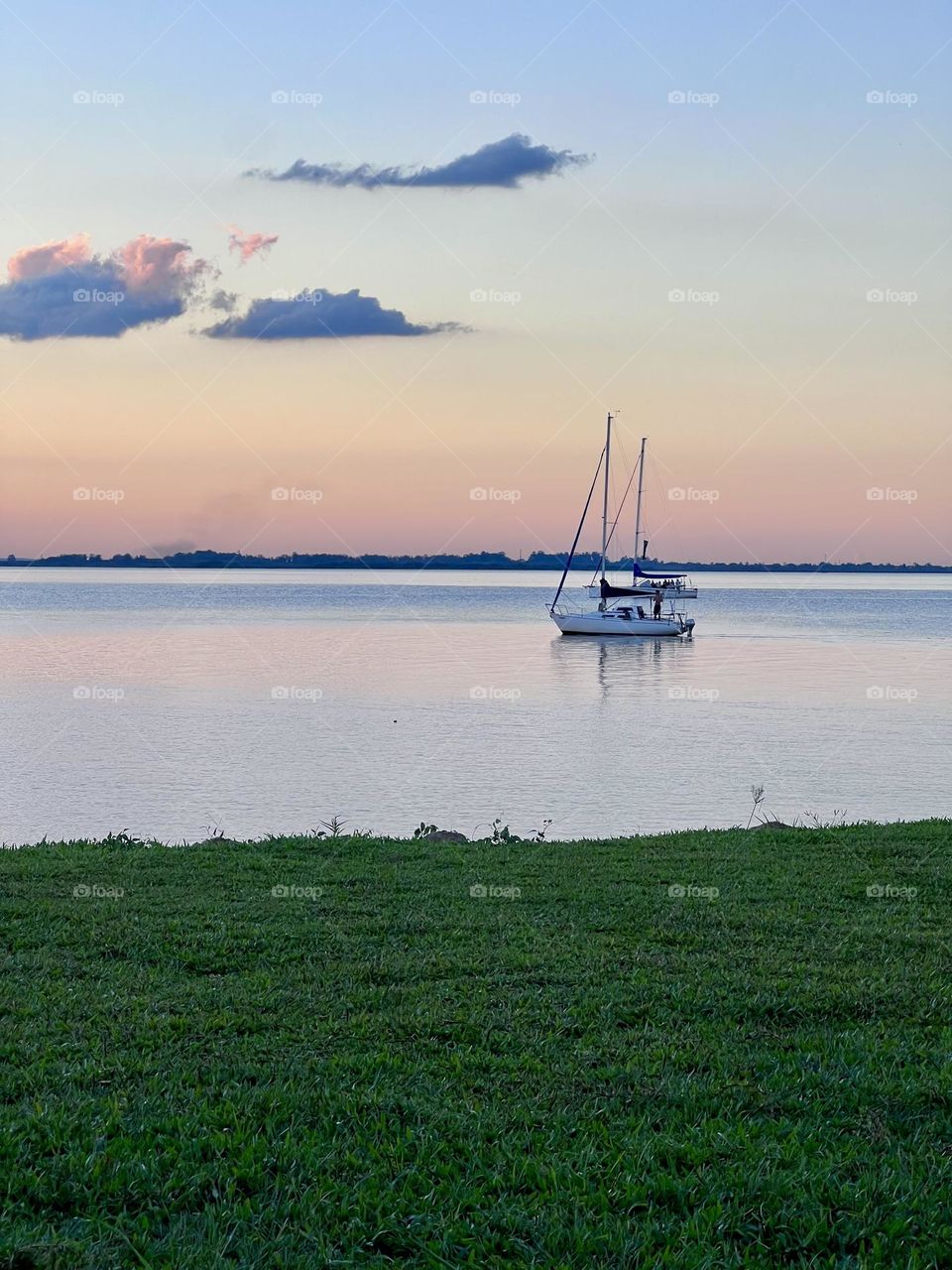Quiet scene of a sailboat in a quiet lake at sunset, with sky in pastel tones and soft green grass in the foreground, transmitting relaxation and tranquility.