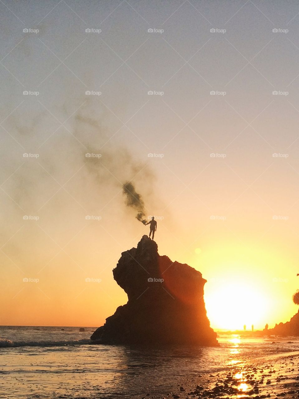 Guy standing on a rock in the ocean holding a smoke stick at sunset. 