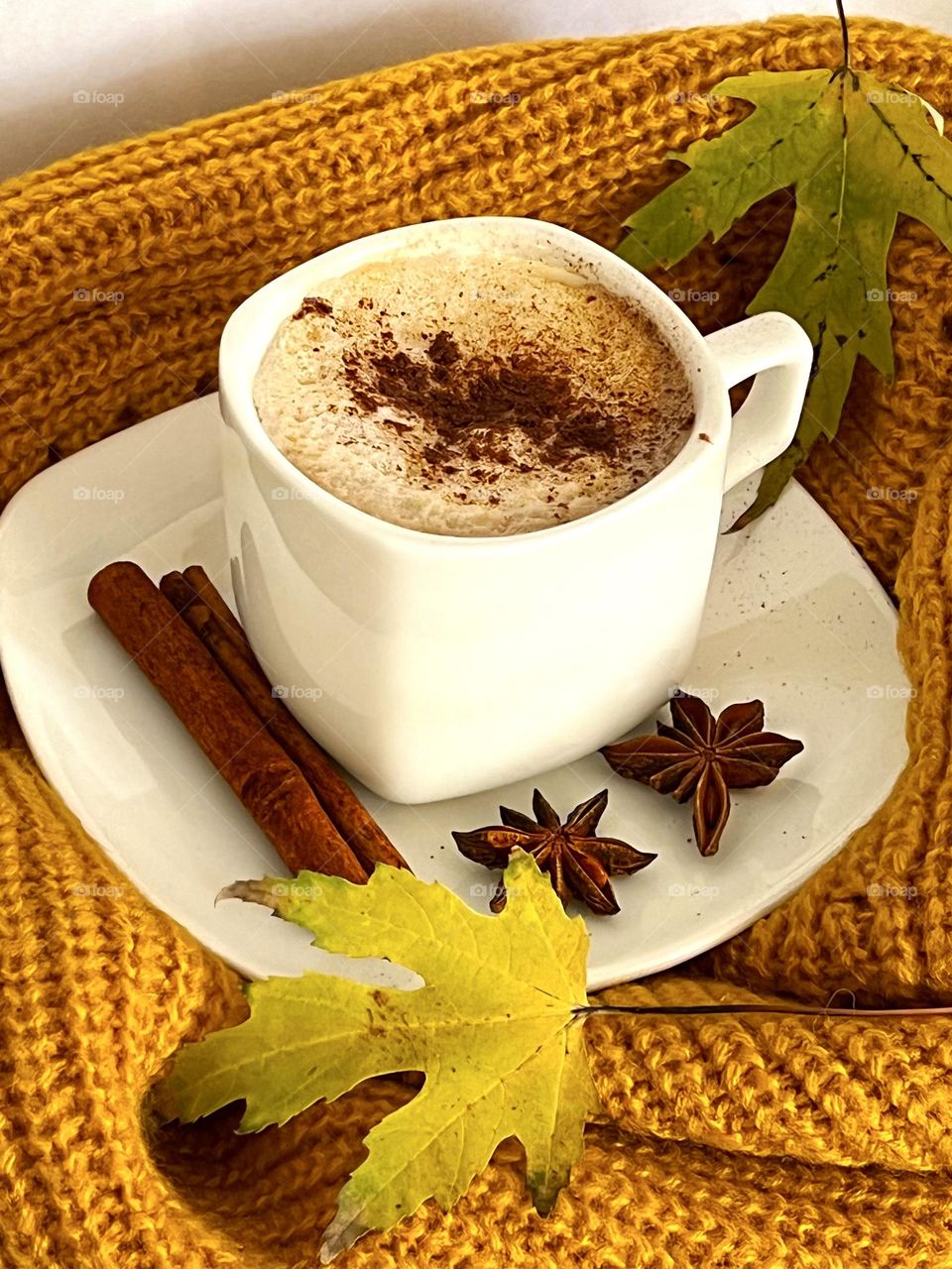White cup of coffee with cinnamon, star anise and cinnamon on the saucer. The cup stands on an orange knitted scarf on a white background. Yellow leaves around