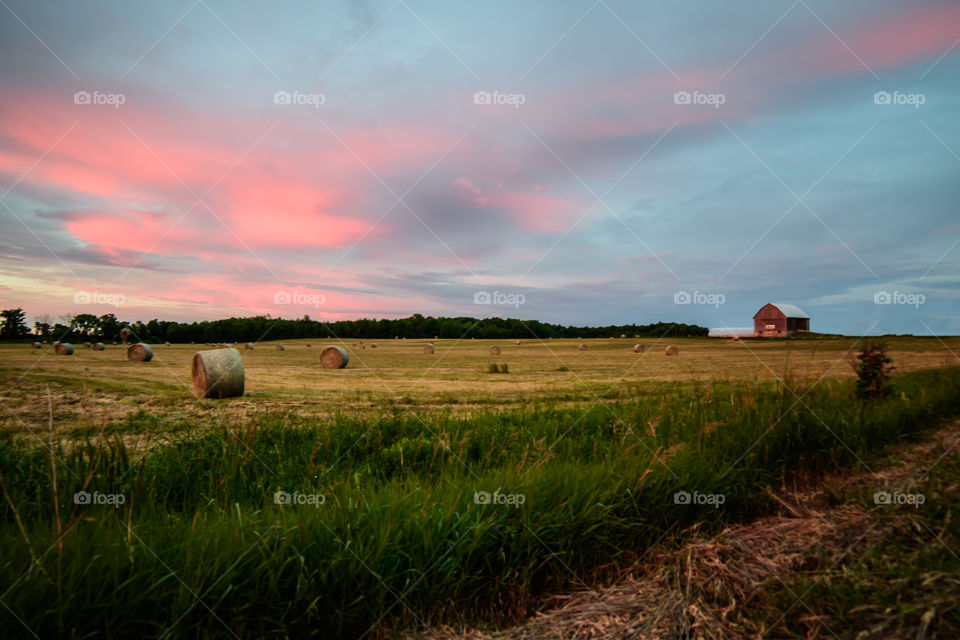 Beautiful sunset on a country road over field with hay and barn in the distance with a pink summer sky 