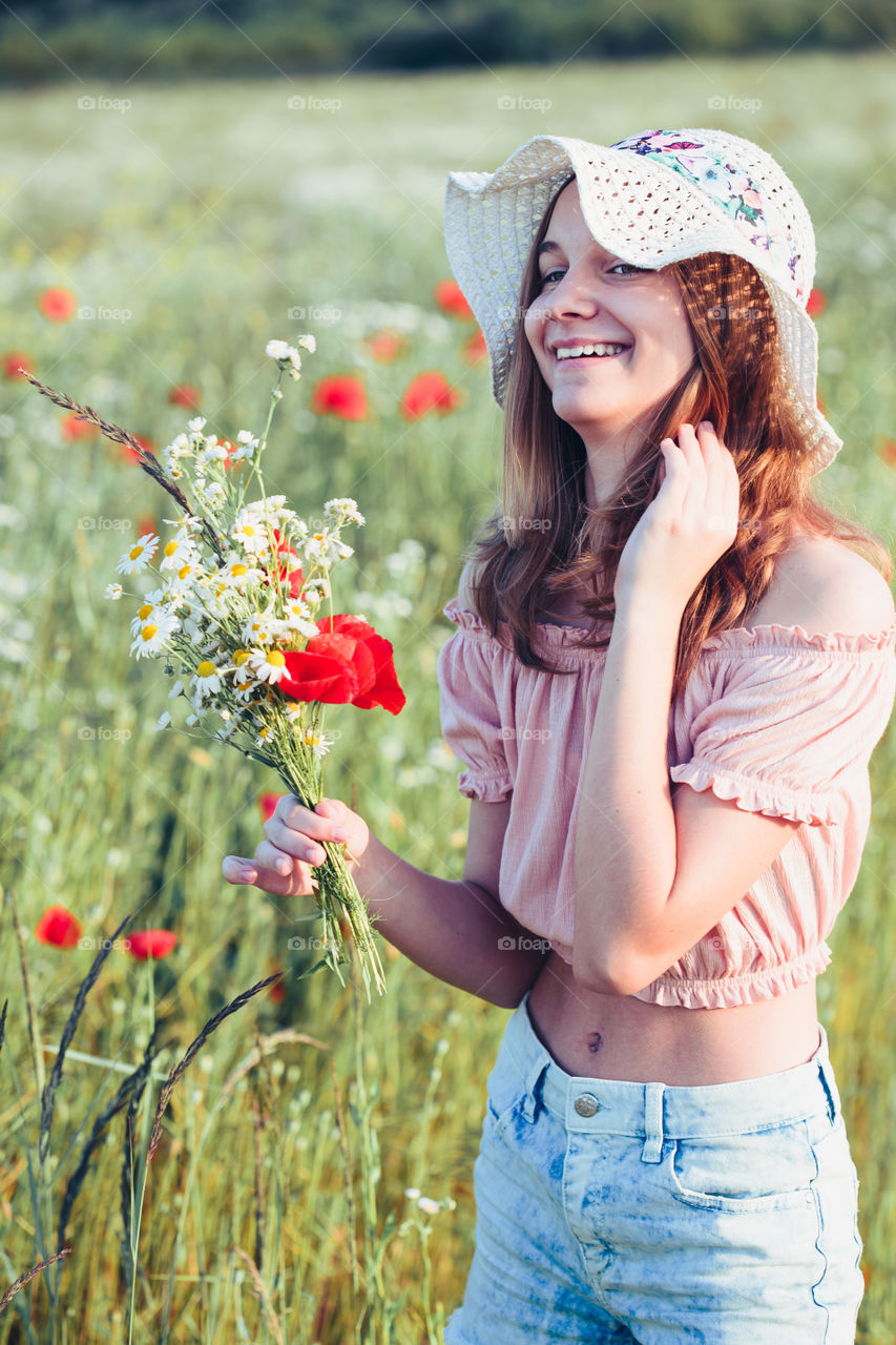 Beautieful young girl in the field of wild flowers. Teenage girl picking the spring flowers in the meadow, holding bouquet of flowers. She wearing hat and summer clothes. Spending time close to nature