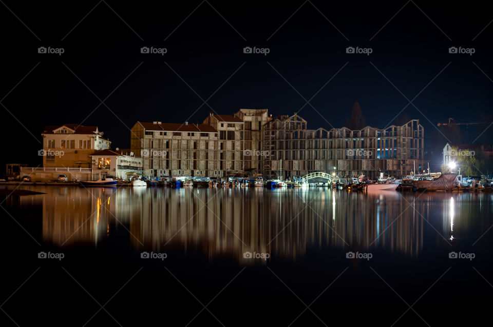 Night city landscape, in the smooth surface of sea water an unfinished building is reflected in the light of lanterns