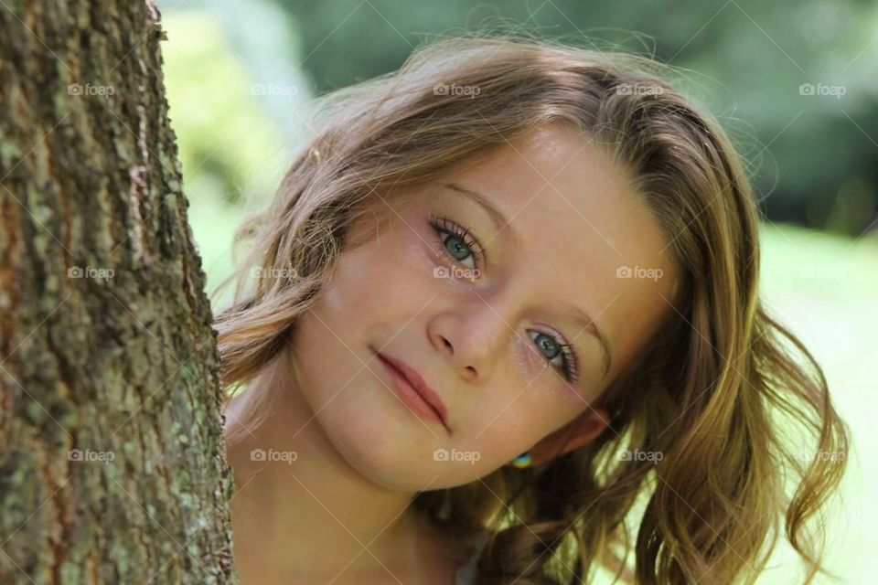 Young Girl with Big blue eyes and brown hair, peeking around a tree outdoors
