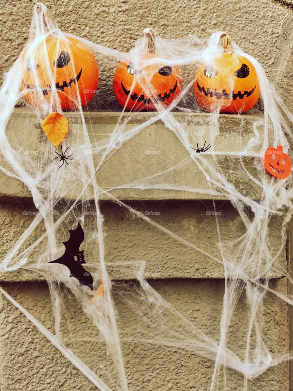 Halloween. Three scary pumpkins lie on the parapet of a stone wall and are covered with cobwebs. Hanging on the web: bat, spider yellow autumn leaves
