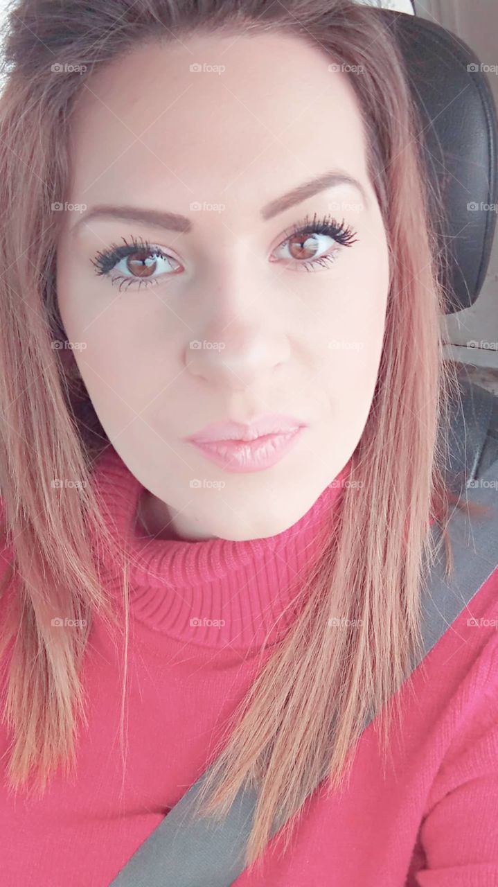 Young girl sitting in a car, brown hair, light makeup. Her brown eyes stare back at her phone as she takes a cute, but compact selfie.